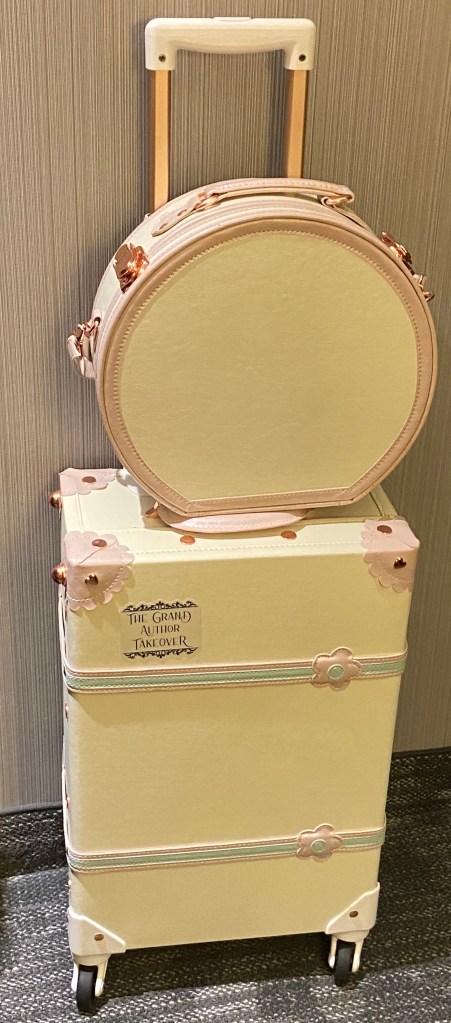 Image of a cream-coloured wheeled suitcase with pink and blue trim, rose-gold hardware. A matching round hatbox-style case sits on top of it. The suitcase has a decal on the front from "The Grand Author Takeover"